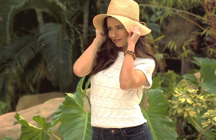 Woman in a white sweater and hat posing outdoors, embodying celebrity style amid lush greenery.