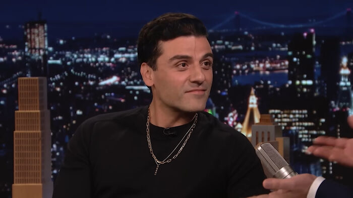 Celebrity on a talk show, wearing a black shirt and a gold chain, engaging with the host against a cityscape backdrop.