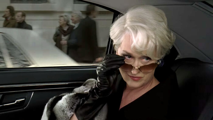 Actor in car wearing sunglasses and black gloves, epitomizing a specific role with an iconic gaze.