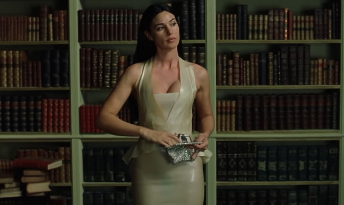 Celebrity in a stylish dress standing in front of a bookshelf, holding a sparkling clutch.