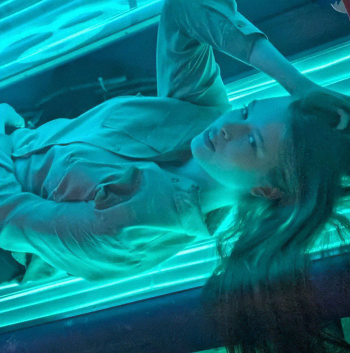 Woman under bright teal lights, lying down and looking upwards, reflecting an eerie atmosphere. Woman under bright teal lights, lying down and looking upwards, reflecting an eerie atmosphere.