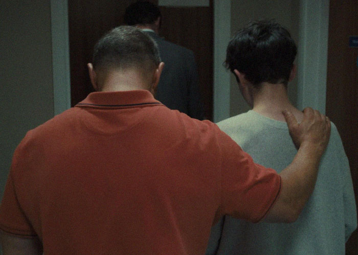 Two people walking down a hallway, one in an orange shirt, related to TV show "Adolescence" and emoji meanings. Two people walking down a hallway, one in an orange shirt, related to TV show "Adolescence" and emoji meanings.