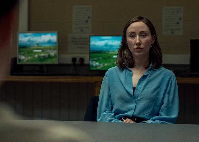 Woman in a blue shirt appearing serious, sitting in front of computers, related to TV show "Adolescence" and emoji meanings. Woman in a blue shirt appearing serious, sitting in front of computers, related to TV show "Adolescence" and emoji meanings.