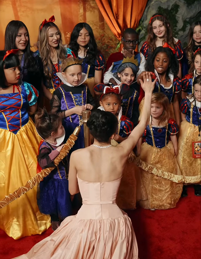 A group of children in Snow White costumes interacting with a woman in an elegant dress on a red carpet. A group of children in Snow White costumes interacting with a woman in an elegant dress on a red carpet.