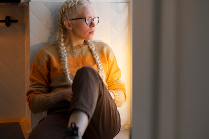 Woman with braids and glasses in a thoughtful pose, sitting by a wall in warm light. Woman with braids and glasses in a thoughtful pose, sitting by a wall in warm light.