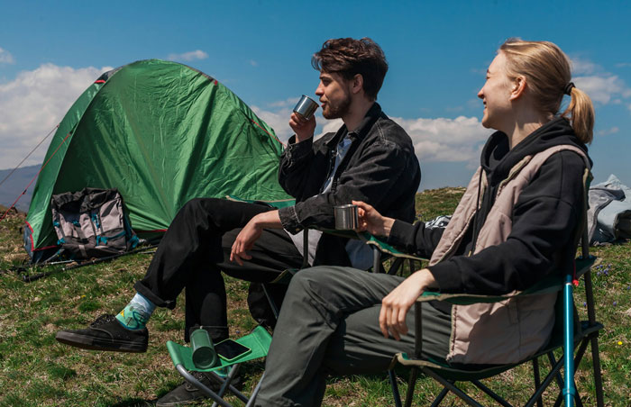 Two people sitting outside a green tent enjoying drinks in a campsite under a blue sky. Two people sitting outside a green tent enjoying drinks in a campsite under a blue sky.