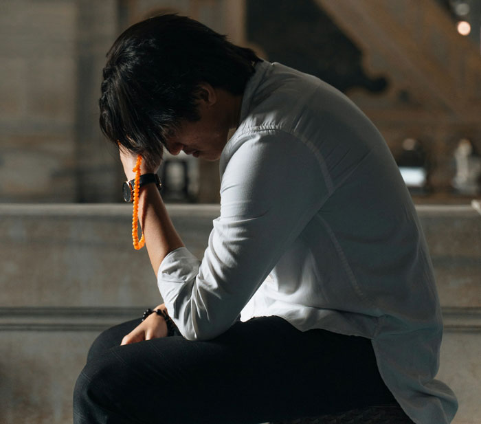 Person in deep thought, seated indoors, wearing white shirt and holding prayer beads, reflecting on family and wealth issues. Person in deep thought, seated indoors, wearing white shirt and holding prayer beads, reflecting on family and wealth issues.