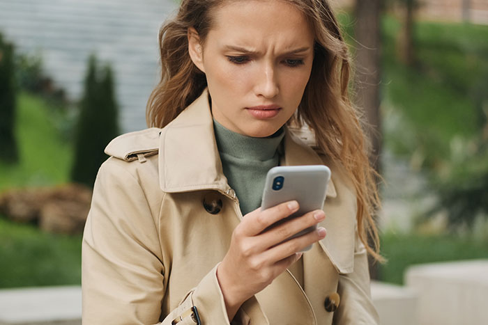 Woman in a trench coat looking concerned at her phone in a park setting. Woman in a trench coat looking concerned at her phone in a park setting.