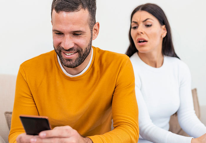 Man and woman sitting on a couch; the woman looks shocked as the man smiles at his phone, creating neighborly tension. Man and woman sitting on a couch; the woman looks shocked as the man smiles at his phone, creating neighborly tension.