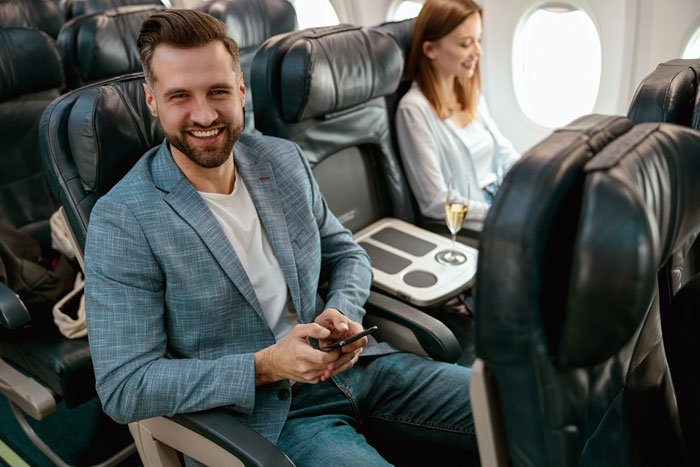 Man smiling while seated on an airplane, holding a smartphone, related to drama about a handicapped seat claim. Man smiling while seated on an airplane, holding a smartphone, related to drama about a handicapped seat claim.