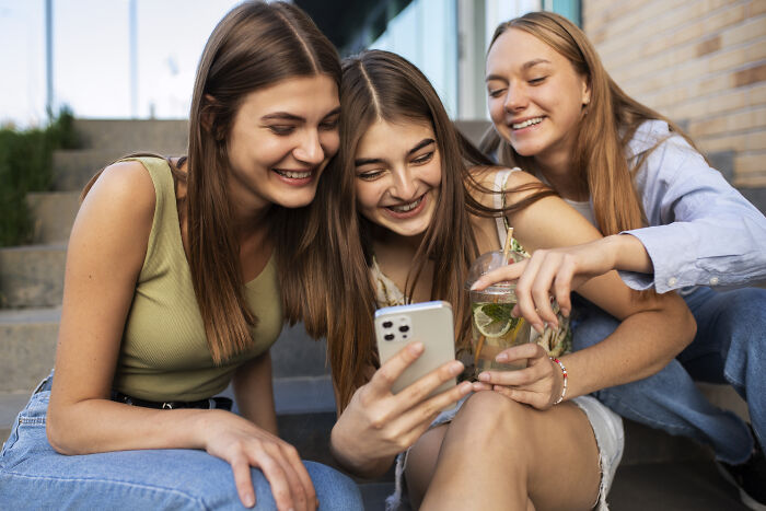 Three people smiling and looking at a smartphone together, sitting on outdoor steps, showcasing a normal societal behavior.