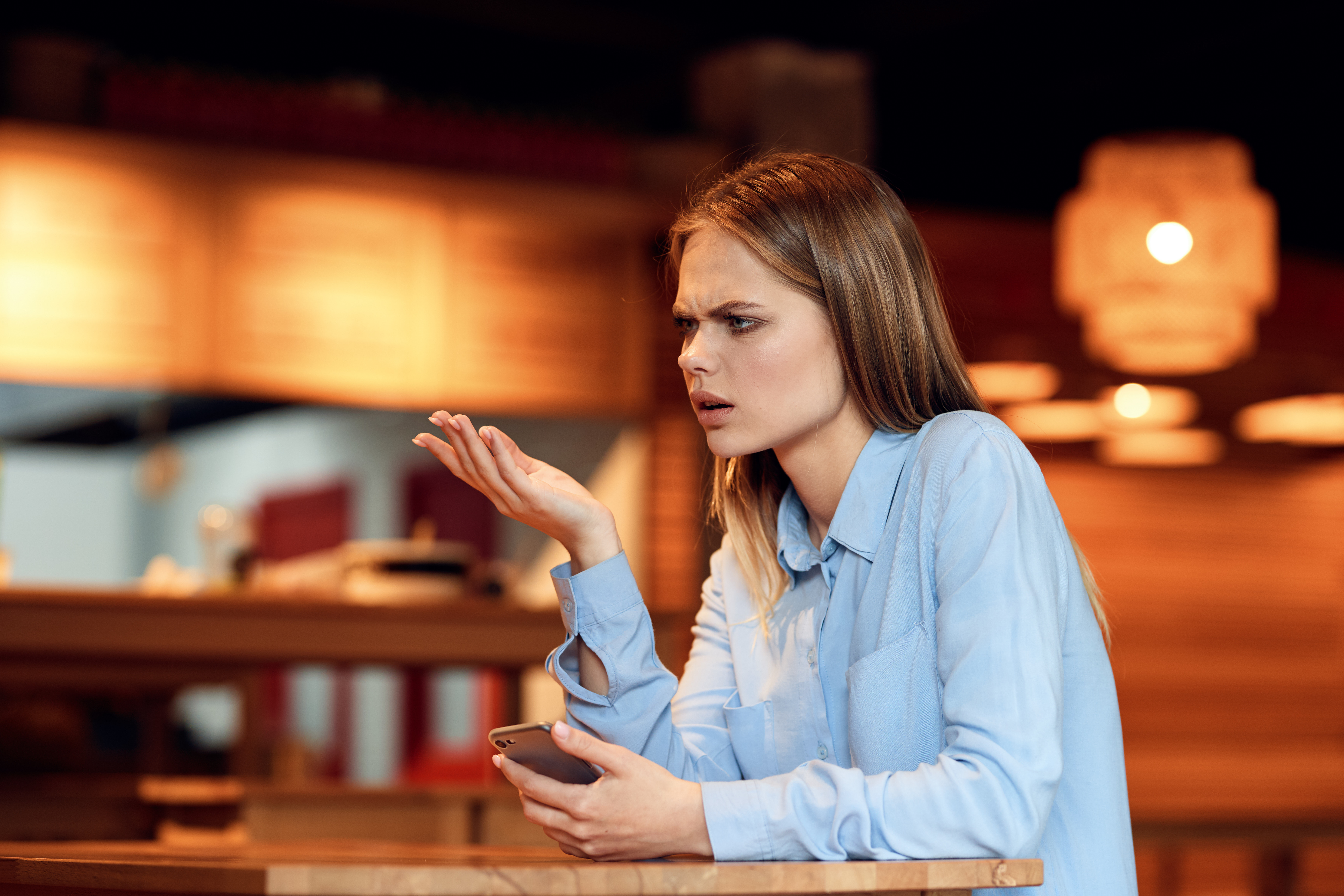 Confused woman in a blue shirt holding a phone at a table, under warm lighting. Confused woman in a blue shirt holding a phone at a table, under warm lighting.