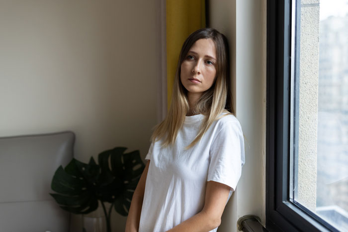 Woman in a white shirt leaning by a window, looking thoughtful. Woman in a white shirt leaning by a window, looking thoughtful.