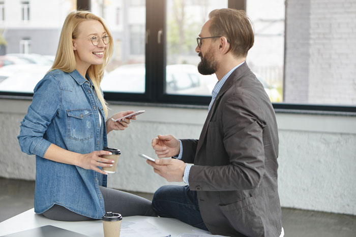 Woman chatting with a handsome man in an office, holding coffee and phones. Woman chatting with a handsome man in an office, holding coffee and phones.