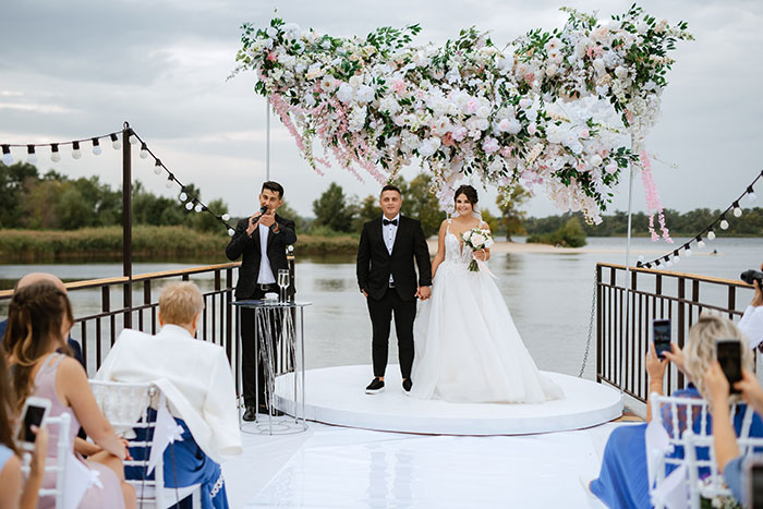 Couple stands at outdoor wedding ceremony under floral arch by a lake, witnessed by seated guests. Couple stands at outdoor wedding ceremony under floral arch by a lake, witnessed by seated guests.