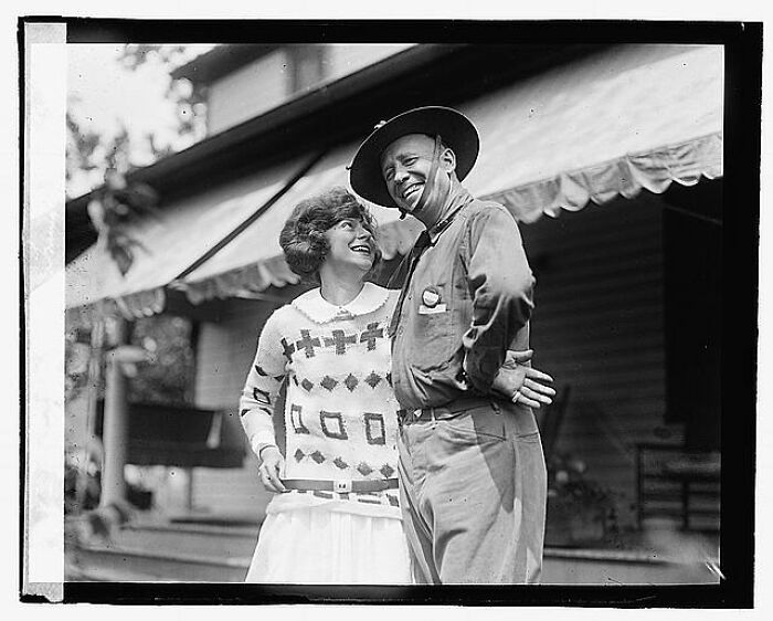 Black and white vintage photo of a smiling couple outside a house, one wearing a patterned sweater and the other a hat.