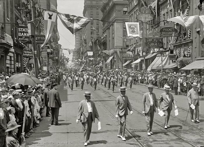 Crowd watching a parade on a busy street showing life in America from 100 years ago with flags and period clothing.
