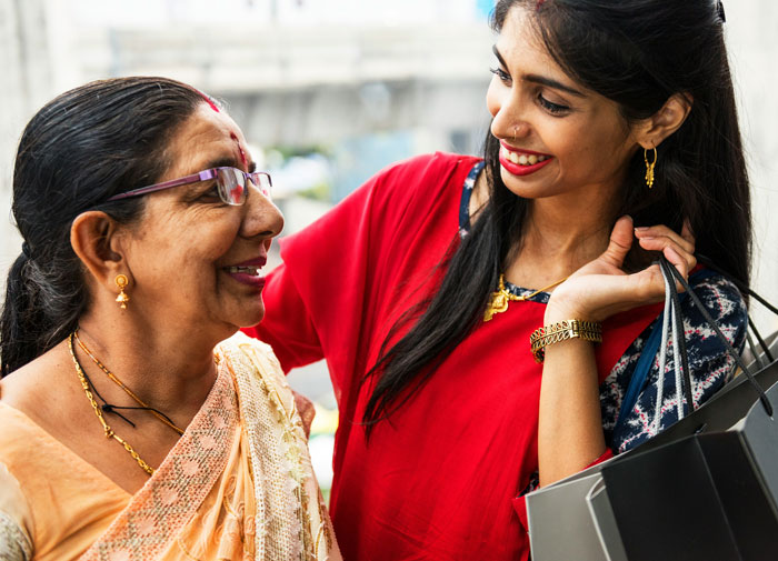 Woman in red smiling at older woman with glasses and a saree, holding shopping bags, outdoors. Woman in red smiling at older woman with glasses and a saree, holding shopping bags, outdoors.