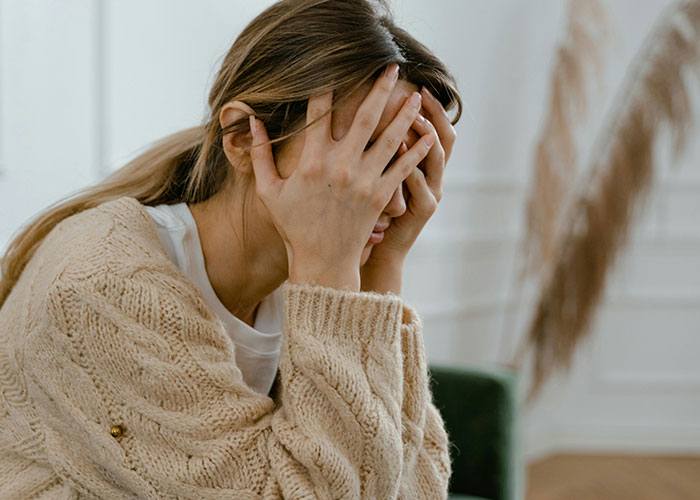 Woman in a beige sweater sitting with face in hands, appearing stressed, in a bright room with decorative plants. Woman in a beige sweater sitting with face in hands, appearing stressed, in a bright room with decorative plants.