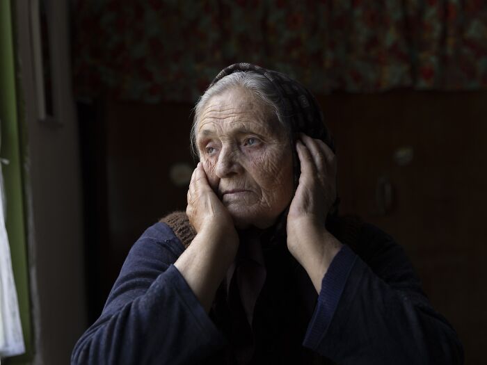 Elderly woman sitting by a window, hands on face, conveying the vulnerability of aging women, by Michele Zousmer.