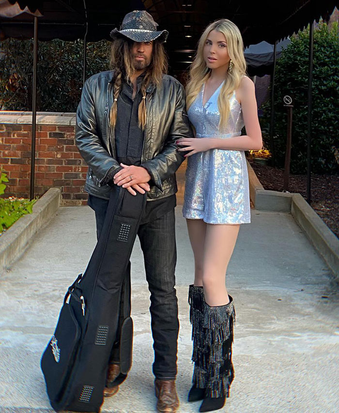 Stylish duo outside, woman in silver dress, man holding guitar case, sparking online discussions about romance on Easter. Stylish duo outside, woman in silver dress, man holding guitar case, sparking online discussions about romance on Easter.