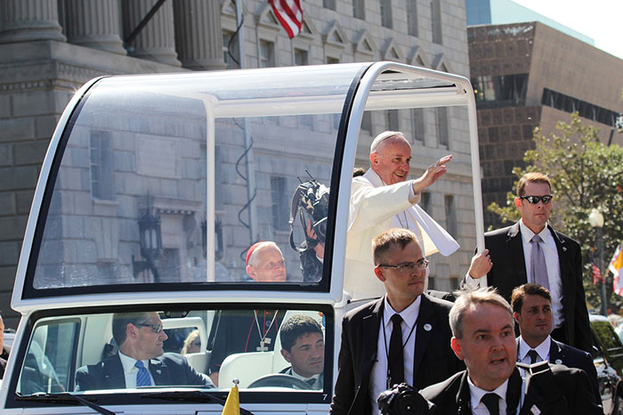 Pope Francis standing in an open popemobile, waving to a crowd, escorted by security personnel. Pope Francis standing in an open popemobile, waving to a crowd, escorted by security personnel.