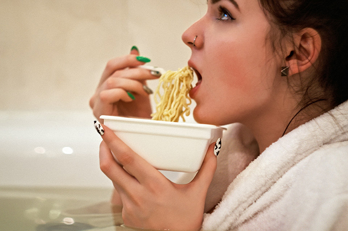 Woman eating noodles in a bathtub, highlighting a gluten-free meal setting. Woman eating noodles in a bathtub, highlighting a gluten-free meal setting.