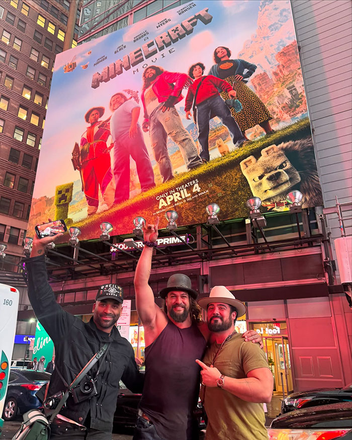 Three people pose under a large "Minecraft" movie poster in a city setting at night. Three people pose under a large "Minecraft" movie poster in a city setting at night.
