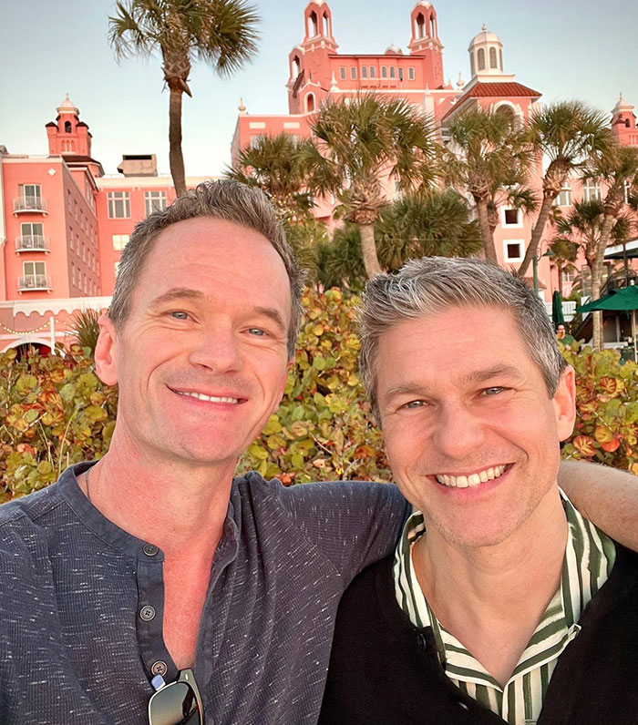 Two smiling men posing in front of a pink building with palm trees, enjoying a sunny day. Two smiling men posing in front of a pink building with palm trees, enjoying a sunny day.