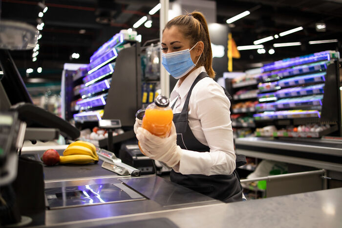 Cashier in a supermarket wearing a mask and gloves, scanning items, illustrating American grocery shopping habits.