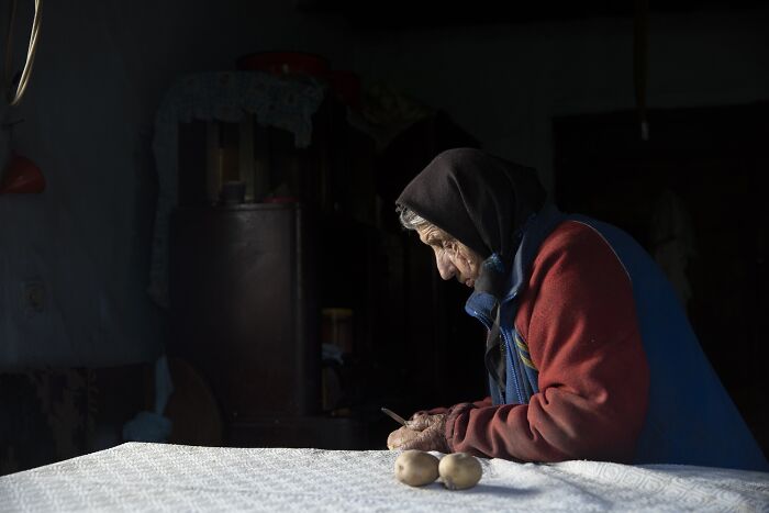 Elderly woman in a dark room, wearing a hooded jacket, peeling potatoes.