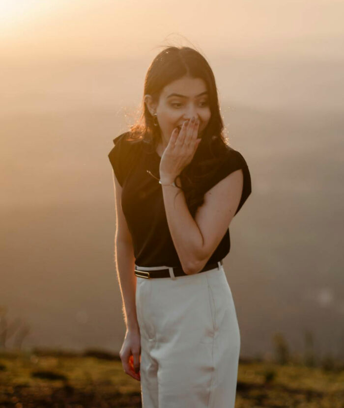Woman in a black top and white pants covering her mouth, expressing embarrassment at sunset.