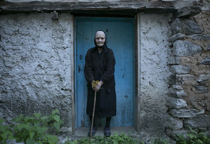 Elderly woman in black stands at a blue door, holding a cane, illustrating the vulnerability of aging women.