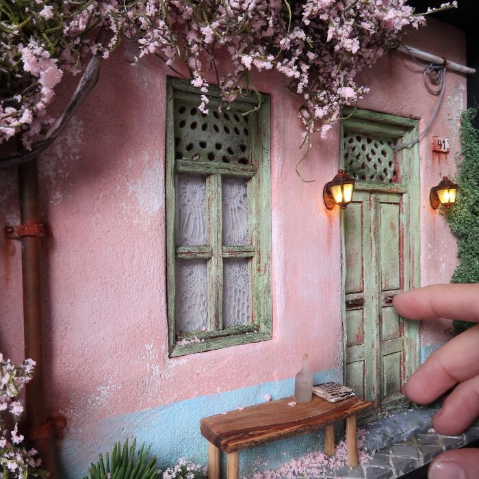Miniature house by Tamar Cohen with pink walls, green doors, and flowers, featuring a bench and hand for scale.