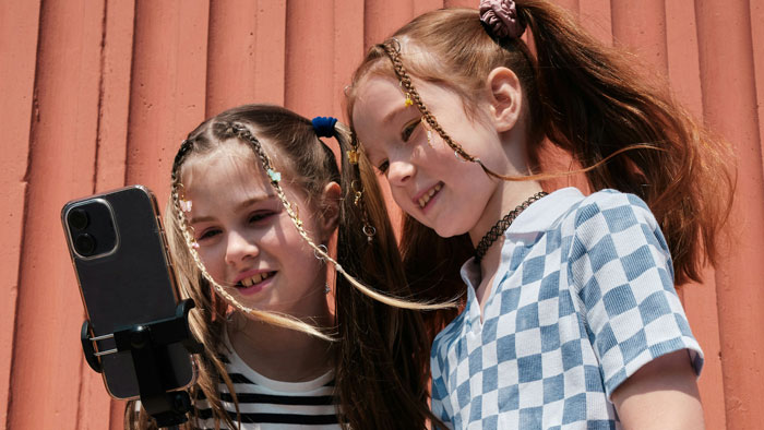 Two young girls smiling at a smartphone on a tripod against a red wall, one with checkered dress and braids, focused on TikTok. Two young girls smiling at a smartphone on a tripod against a red wall, one with checkered dress and braids, focused on TikTok.