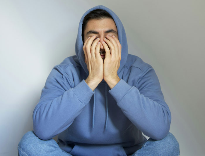Man in blue hoodie sitting with hands on face, expressing stress about video games. Man in blue hoodie sitting with hands on face, expressing stress about video games.