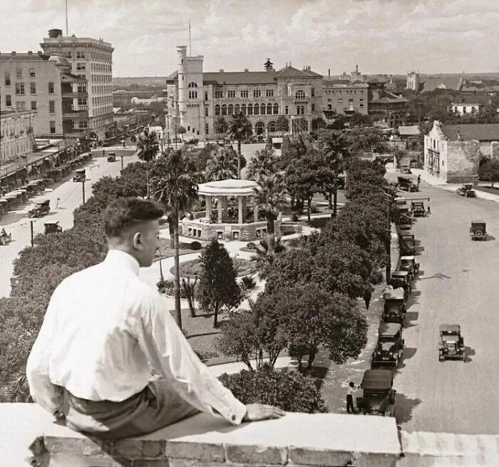 Young man sitting on rooftop ledge overlooking vintage cars and a city park, showing life in America 100 years ago.