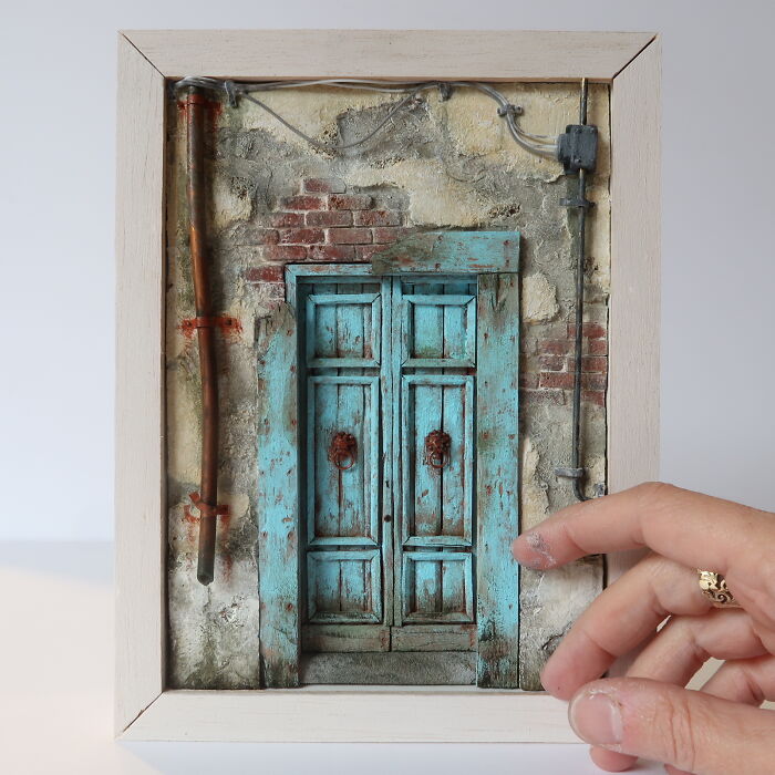 Miniature art by Tamar Cohen featuring a hand adjusting a tiny blue door on a rustic wall.