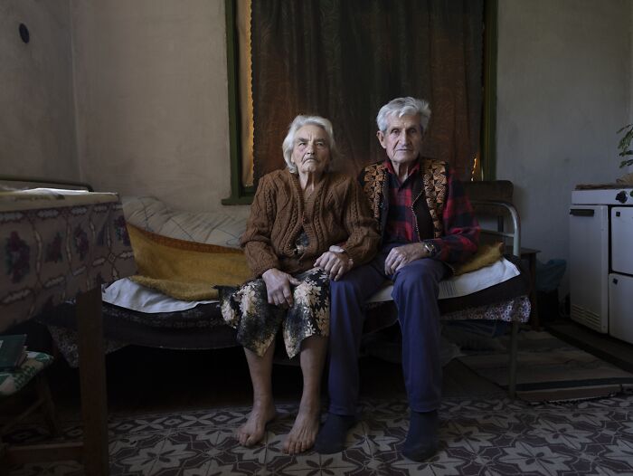 Elderly couple sitting on a bed in a modest room, conveying the vulnerability of aging women.