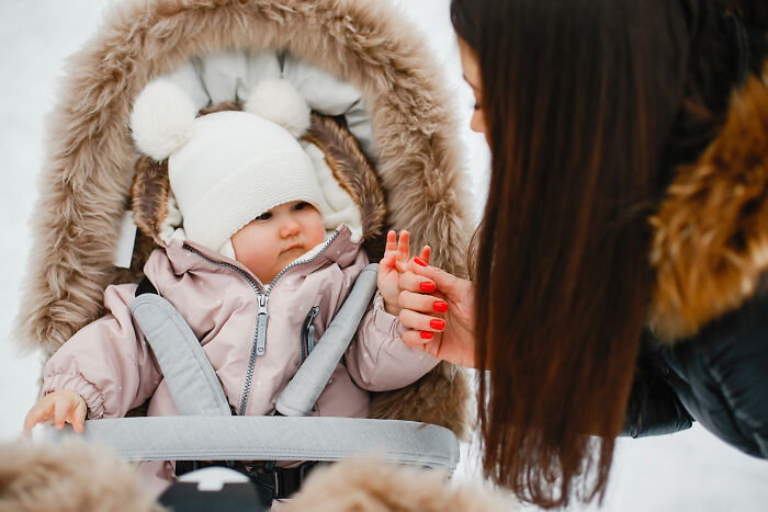 Baby bundled in winter clothes in stroller with fur lining, holding mother's hand in a super normal thing country setting.