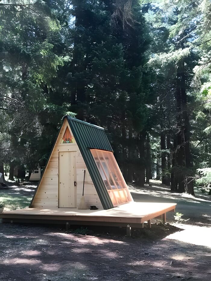 A beautifully crafted woodworking project of an A-frame wooden cabin surrounded by tall forest trees in sunlight.