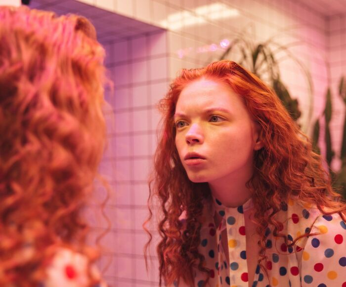 Young woman with red hair and a polka dot shirt gazing intently in a mirror, showcasing skillful expression.