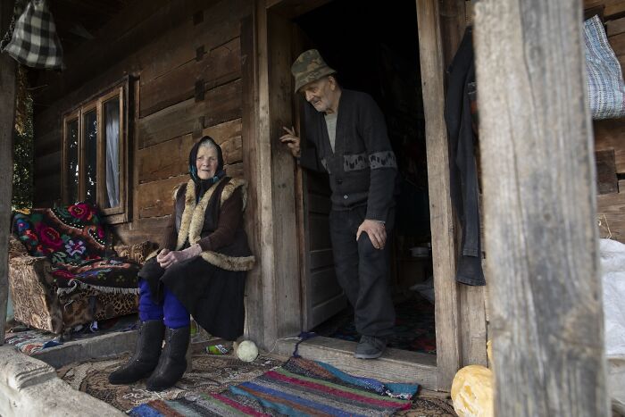 An elderly woman sitting on a porch conveying the vulnerability of aging, as a man stands nearby in a rustic setting.
