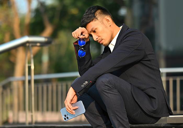 Man in suit sitting outdoors, looking contemplative, holding a phone and sunglasses, related to wedding and plus one issue. Man in suit sitting outdoors, looking contemplative, holding a phone and sunglasses, related to wedding and plus one issue.