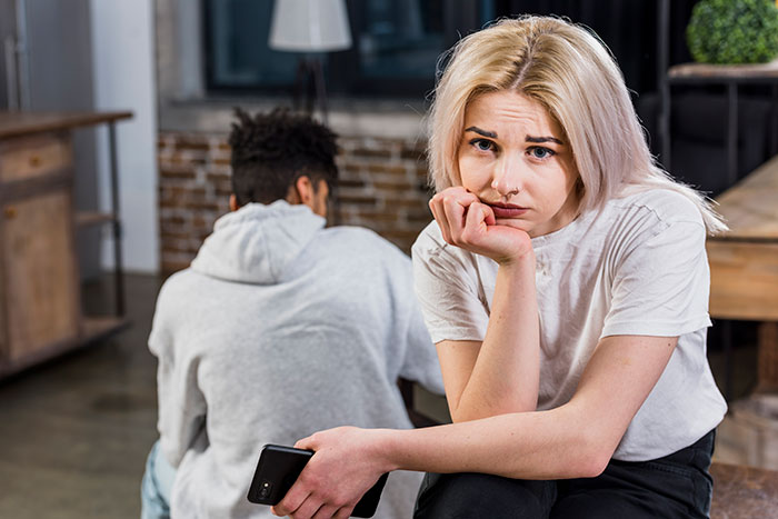 Woman looking upset with her partner turned away, holding a phone; relationship tension visible. Woman looking upset with her partner turned away, holding a phone; relationship tension visible.