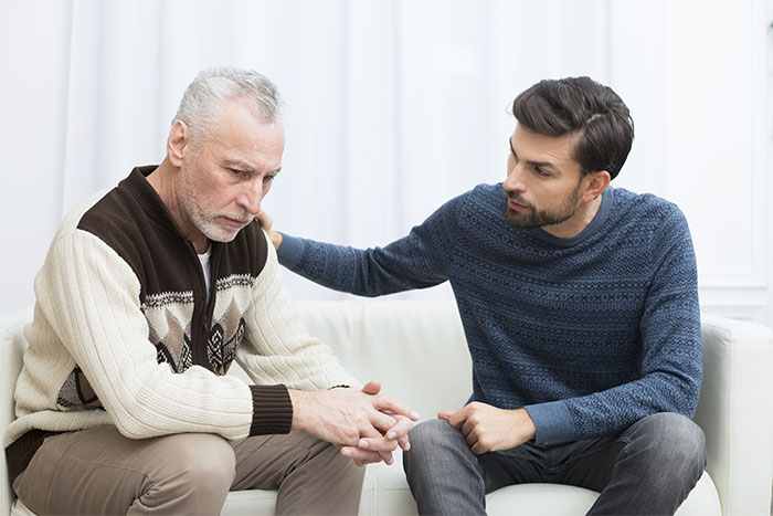 Two men sitting on a couch in conversation, one comforting the other, discussing a banned babysitting incident. Two men sitting on a couch in conversation, one comforting the other, discussing a banned babysitting incident.