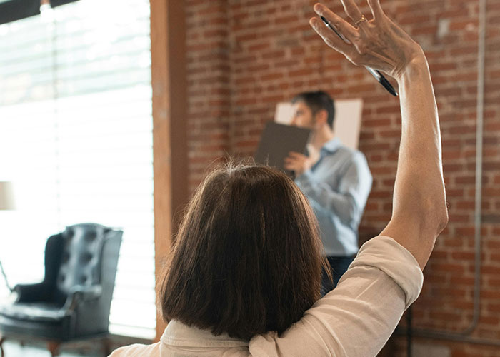 A woman raising her hand in a classroom setting with a speaker in the background, brick wall visible. A woman raising her hand in a classroom setting with a speaker in the background, brick wall visible.