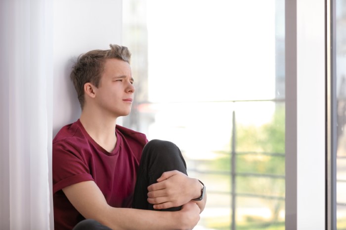 Teen sitting by a window, looking distressed, wearing a maroon shirt, contemplating harassment issues. Teen sitting by a window, looking distressed, wearing a maroon shirt, contemplating harassment issues.