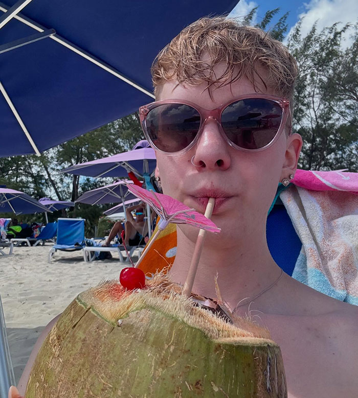 A person on the beach sipping a drink from a coconut under an umbrella, wearing pink sunglasses. A person on the beach sipping a drink from a coconut under an umbrella, wearing pink sunglasses.