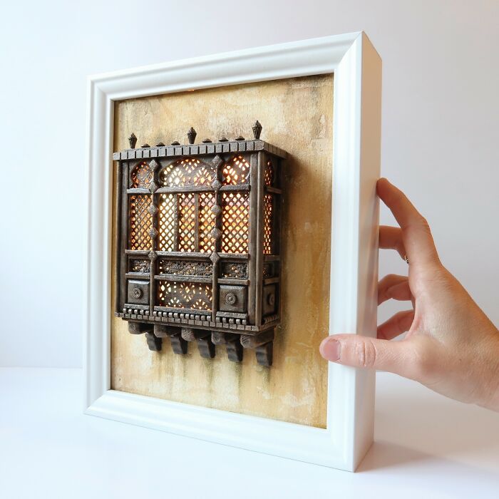 A hand holding a frame with a detailed, illuminated miniature depicting a tiny world by Tamar Cohen.